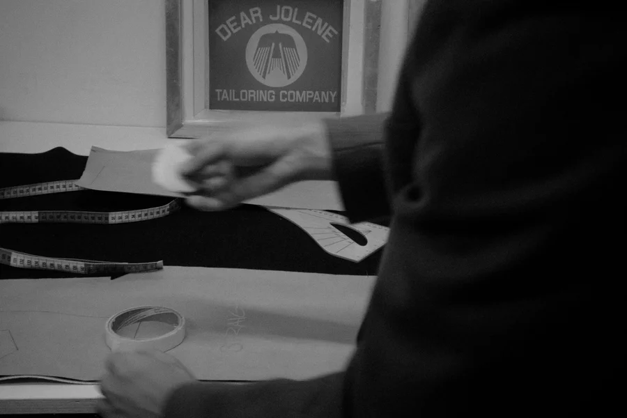 A tailor marking a paper pattern on dark cloth at a workbench, below a framed Dear Jolene Tailoring Company sign.