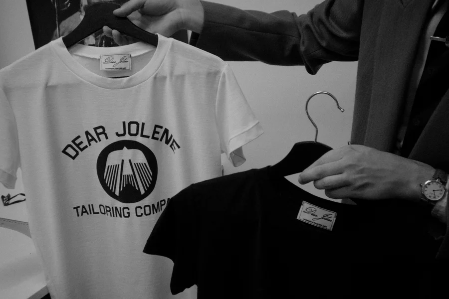 Two Dear Jolene Tailoring Company T-shirts on hangers — a white logo tee and a black tee with a woven label.